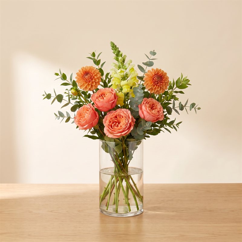 Coral garden roses, orange dahlias and yellow snapdragons with eucalyptus in a clear vase